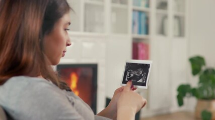 Wall Mural - Beautiful pregnant woman with brown hair stitting in front of cosy fireplace stroking baby belly look at ultrasound pictures shot in 4k