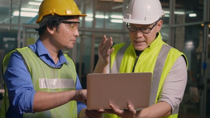 Two factory industrial workers technician or engineer and manager wear uniforms safety feeling upset with the engine machine of the factory arguing about planning procedure of work on laptop computer