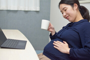 A young pregnant Asian woman cares her baby in her stomach and drinks a tea during telework