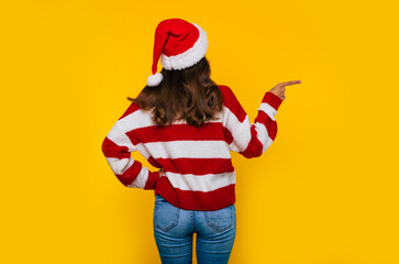 Wall Mural - Back view photo of a young woman in Christmas hat and striped winter sweater is pointing away while she isolated on yellow background