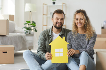 Portrait of happy young married couple sitting on floor in living room interior in new home, holding symbolic paper house, looking at camera and smiling. Real estate, mortgage, buying property concept