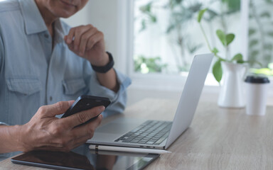 Canvas Print - Asian business man using mobile phone, thinking about his work project during working on laptop computer with digital tablet on table at home office