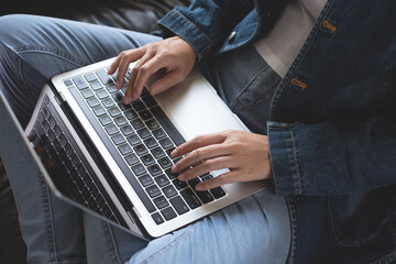 Wall Mural - Young casual woman, freelancer in blue jeans and sweater sitting on sofa working and typing on laptop computer keyboard at home. Student studying online class, E-learning, working from home, close up