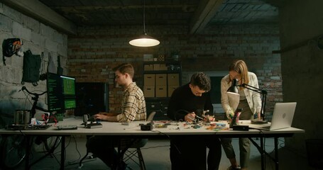 Garage Office with 3 People Team Working on Technology Electronics Developing Startup in Loft Basement Room at Night. Robotics Engineering Design Concept with Soldering and Coding. 4K Wide Pan Video