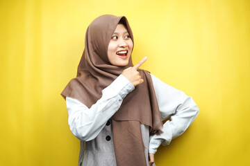 Beautiful young asian muslim woman shocked, surprised, wow expression, with hand pointing to empty space, presenting something, presenting product, isolated on yellow background