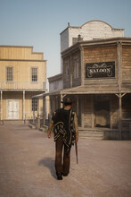 3D Rendering Of A Cowboy Or Gunman Walking Towards A Saloon With A Rifle In His Hand In An Old Wild West Town.