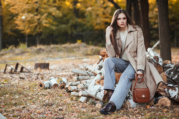 A beautiful tall girl of model appearance in a beige raincoat and gray jeans sits on sawn logs in a city park on a warm autumn day. Autumn street trendy look.