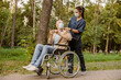 Young nurse in face shield and protective face mask assisting senior handicapped man in wheelchair using his smartphone during a walk in the park