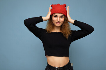 Wall Mural - Photo of young pretty positive brunette woman curly with sincere emotions wearing stylish black crop top and red hat isolated on blue background with copy space