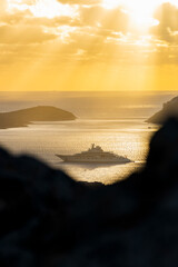 Wall Mural - (Selective focus) Stunning sunrise during a cloudy day with a rock in the foreground and a luxury yacht sailing on a calm water in the distance. Golfo Aranci, Sardinia, Italy.