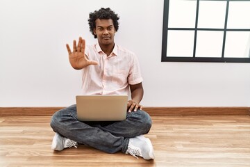 Canvas Print - African man with curly hair using laptop sitting on the floor doing stop sing with palm of the hand. warning expression with negative and serious gesture on the face.