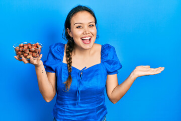 Young hispanic girl holding bowl of chestnuts celebrating achievement with happy smile and winner expression with raised hand