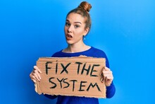 Young Blonde Woman Holding Fix The System Banner Cardboard In Shock Face, Looking Skeptical And Sarcastic, Surprised With Open Mouth