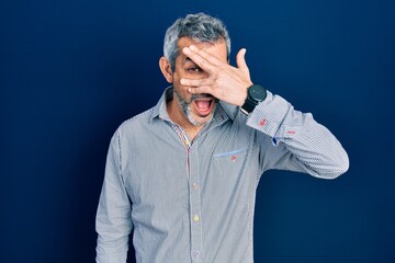 Canvas Print - Handsome middle age man with grey hair wearing business shirt peeking in shock covering face and eyes with hand, looking through fingers with embarrassed expression.