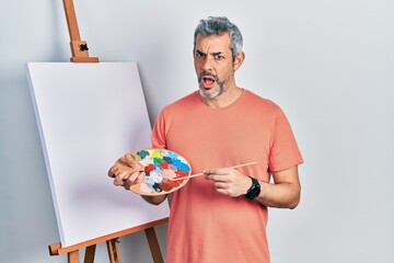 Poster - Handsome middle age man with grey hair standing drawing with palette by painter easel stand in shock face, looking skeptical and sarcastic, surprised with open mouth