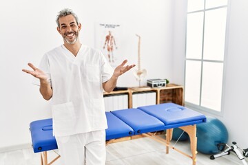 Poster - Middle age hispanic therapist man working at pain recovery clinic smiling cheerful offering hands giving assistance and acceptance.