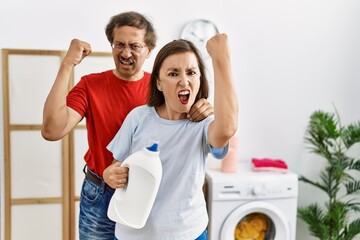 Sticker - Middle age interracial couple doing laundry holding detergent bottle annoyed and frustrated shouting with anger, yelling crazy with anger and hand raised