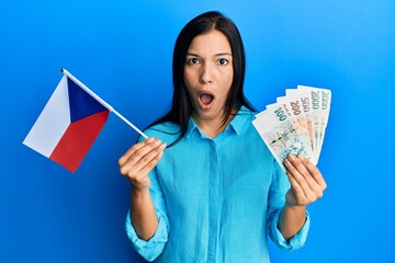 Sticker - Young latin woman holding czech republic flag and koruna banknotes afraid and shocked with surprise and amazed expression, fear and excited face.