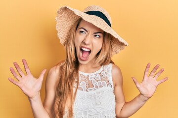 Wall Mural - Young caucasian woman wearing summer hat crazy and mad shouting and yelling with aggressive expression and arms raised. frustration concept.