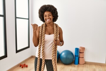 Canvas Print - Young african american woman with afro hair at the gym training with battle ropes pointing to the back behind with hand and thumbs up, smiling confident