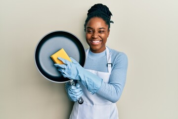 Sticker - African american woman with braided hair wearing apron holding scourer washing pan winking looking at the camera with sexy expression, cheerful and happy face.