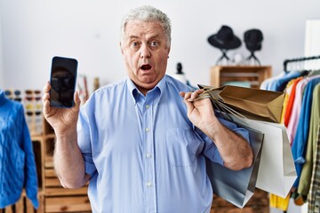 Sticker - Senior man with grey hair holding shopping bags and showing smartphone screen clueless and confused expression. doubt concept.