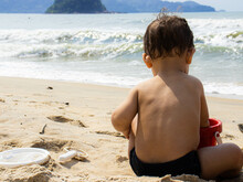 Back Of Child Sitting On The Sand Playing With Toys On The Beach With The Sea In The Background.