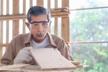 Elderly carpenter wearing safety glasses goggles blowing sawdust off piece of wood plank, senior craftsman working at wooden desk in carpentry woodwork workshop, enjoying his DIY hobby.