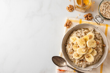 Wall Mural - Banana, walnuts and honey oatmeal porridge in a bowl with a spoon on white table for healthy tasty breakfast. Top view
