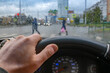 view of the driver hand on the steering wheel of a car against the background of a wet windshield with raindrops and people walking along a pedestrian crossing at an intersection with a traffic light