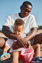 Wall Mural - Vertical portrait of young African-American father and son playing in sand together at beach