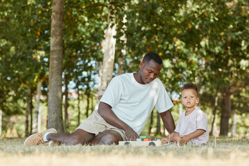 Wall Mural - Portrait of young African-American father playing with cute son in park while sitting on grass, copy space