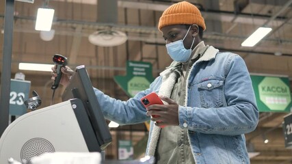 Wall Mural - African American guy in disposable mask types on smartphone standing near self-checkout with large display in store low angle shot