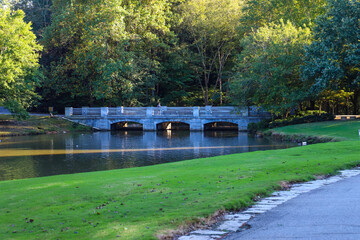 a stone bridge over the lake surrounded by green grass and lush green and autumn colored trees with still silky brown lake water at Lenox Park in Brookhaven Georgia USA