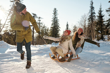 Wall Mural - Two happy girls in winterwear laughing while young man pulling sledge and running