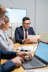 Poster - Serious mature broker looking through financial papers in front of young employees