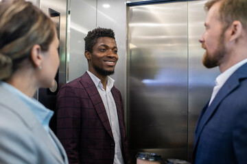 Poster - Happy young African businessman smiling while looking at male colleague during communication inside elevator