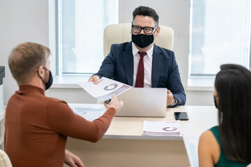 Poster - Mature businessman in protective mask passing financial paper to young man during consultation