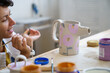 © DimaBerlin - Art therapy: happy smiling woman hold paintbrush looking at pottery jug with cute hand drawn ornament. Female enjoy visiting artistic classes and ceramic workshop studio. Craftswoman busy with work