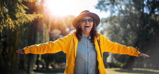 Beautiful happy senior mature woman in hat and yellow jacket enjoying nature in the park stretching her arms