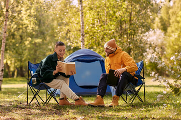 Canvas Print - camping, tourism and travel concept - happy couple with tablet pc computer and book resting at tent camp