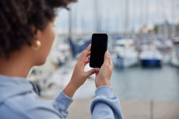 Unrecognizable woman takes photo of harbor poses on pier holds modern mobile phone with blank screen admires sunset goes sailing dressed casually. People tourism travel and spare time concept