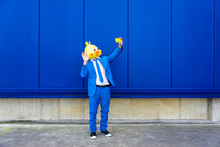 Man Wearing Vibrant Blue Suit And Bird Mask Taking Smart Phone Selfie In Front Of Blue Wall