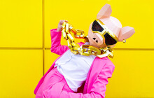 Woman Wearing Vibrant Pink Suit, Pig Mask And Large Golden Chain Posing Against Yellow Wall