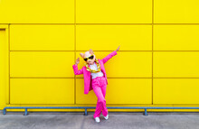 Woman Wearing Vibrant Pink Suit, Pig Mask And Large Golden Chain Posing With Raised Arms In Front Of Yellow Wall