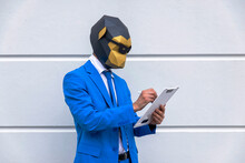 Man Wearing Vibrant Blue Suit And Monkey Mask Writing In Clipboard In Front Of White Wall
