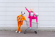 Man And Woman Wearing Vibrant Suits And Animal Masks posing With Shopping Cart And Disco Ball In Front Of White Wall