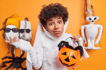 Wall Mural - Hesitant clueless young Afro American woman shrugs shoulders looks unaware holds carved pumpkin and spider thinks about halloween celebration in family circle poses against orange background
