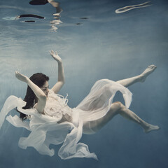 Wall Mural - A girl with dark hair in a white dress swims underwater as if flying in zero gravity