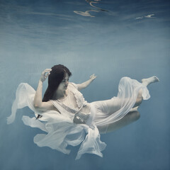 A girl with dark hair in a white dress swims underwater as if flying in zero gravity